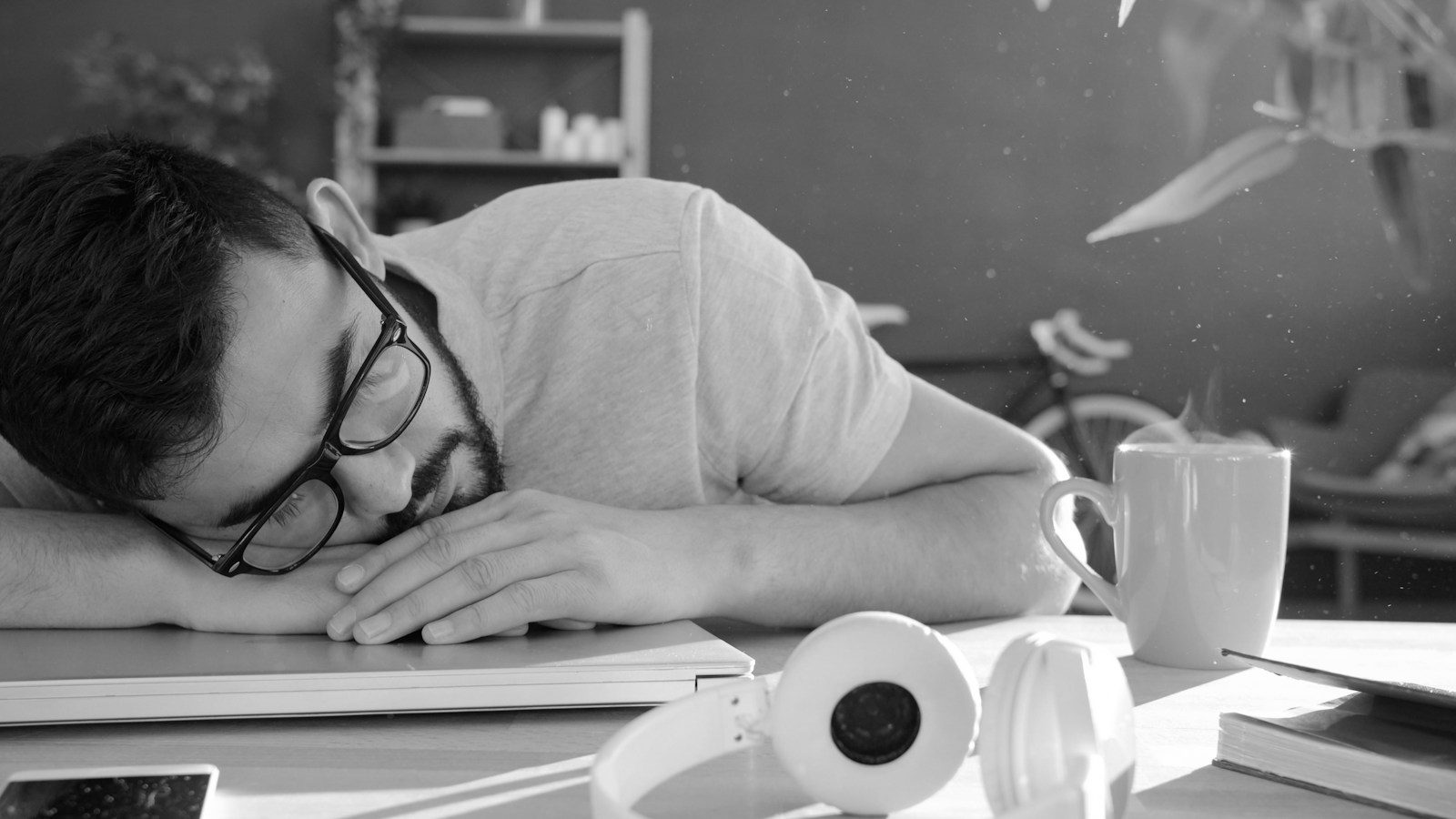 Un hombre duerme con la cabeza sobre un portátil cerrado, junto a unos audífonos y una taza de café